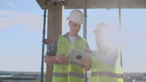 Ein-Bauarbeiter-Und-Eine-Architektin-Mit-Helm-Besprechen-Den-Bauplan-Für-Ein-Haus,-Erzählen-Sich-Gegenseitig-Etwas-über-Den-Entwurf,-Halten-Ein-Tablet-In-Der-Hand-Und-Schauen-Sich-Die-Zeichnungen-Auf-Dem-Hintergrund-Der-Sonnenstrahlen-An.